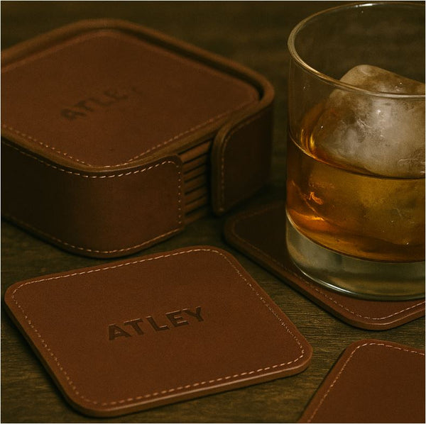 Brown leather coasters with 'ATLEY' branding next to a glass of whiskey on a wooden surface.