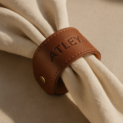 Brown leather napkin ring with 'ATLEY' branding on a beige fabric background