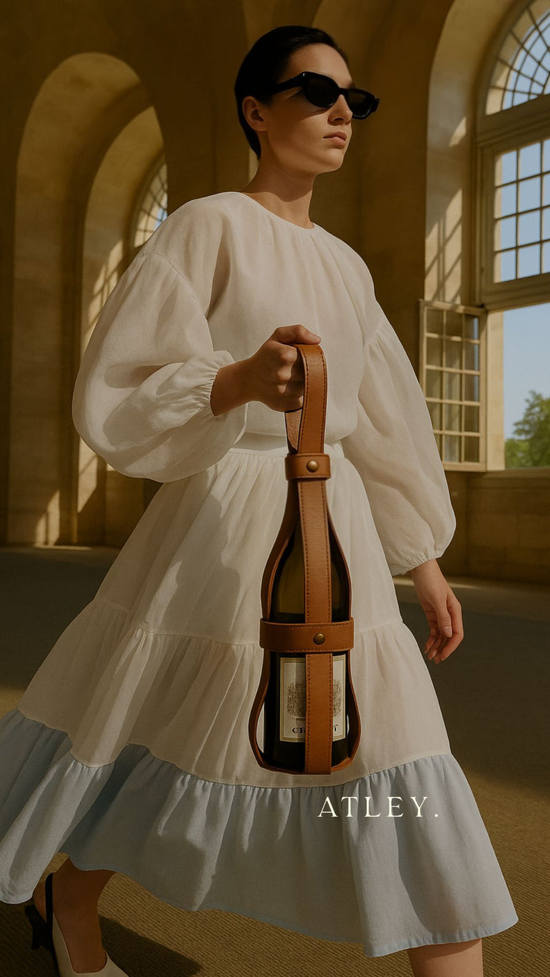 Elegant woman holding a tan leather wine carrier by ATLEY. in a sunlit architectural hallway, wearing a white tiered dress and black sunglasses. Luxury bottle holder designed for 750ml wine bottles